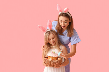 Young woman and her daughter in bunny ears holding basket of Easter cake with painted eggs on pink background - obrazy, fototapety, plakaty