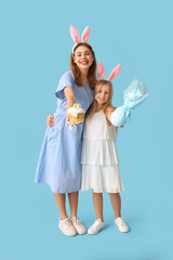 Cute little girl with gift egg and her mother in bunny ears holding Easter cake on blue background