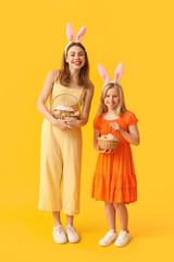 Cute little girl and her mother in bunny ears holding baskets of Easter cake with painted eggs on yellow background