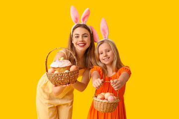 Cute little girl and her mother in bunny ears holding baskets of Easter cake with painted eggs on yellow background