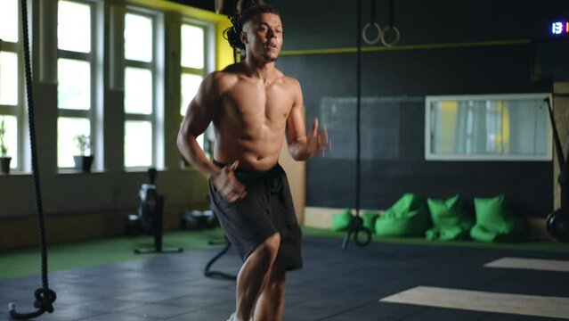 Slow motion. A young mixed race man with dreadlocks on his head performs running in place with a high hip raise in a modern gym