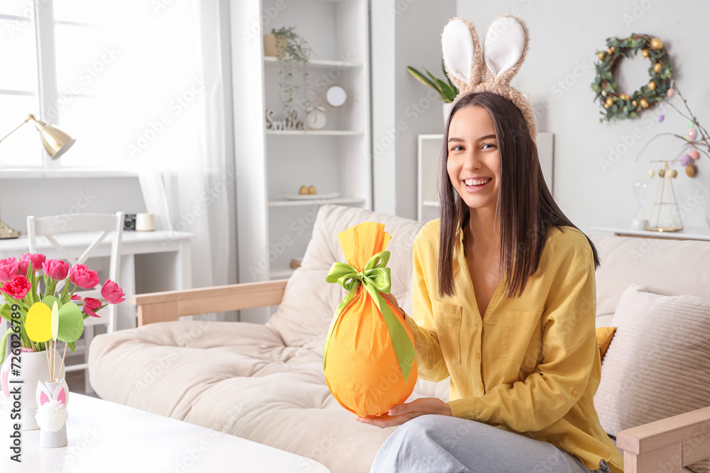 Poster young woman in bunny ears with easter gift egg sitting on sofa at home