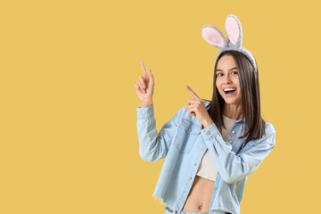 Young woman in bunny ears pointing at something on yellow background. Easter celebration