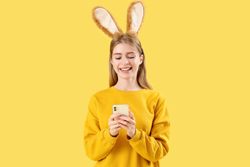Young woman in bunny ears using mobile phone on yellow background. Easter celebration
