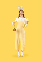 Young woman in bunny ears with basket of Easter eggs on yellow background