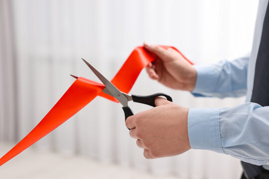 Man cutting red ribbon with scissors indoors, closeup