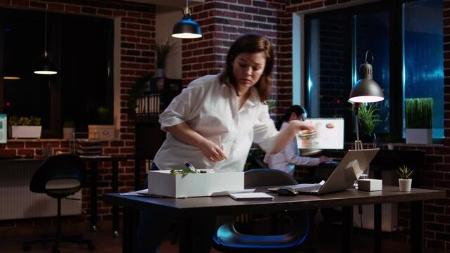 Sad employee leaving office after deciding to quit, waving goodbye to coworker. Upset businesswoman packing desk belongings box in last day in workplace after finishing nightshift, camera A