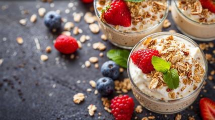 Delicious and healthy greek yogurt in glass cups with oat granola, seeds, berries and mint on a stone counter