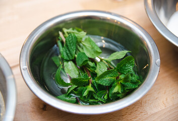 Fresh Mint Leaves in Water Inside Metal Bowl. A close-up of vibrant fresh mint leaves soaking in water in a stainless steel bowl on a wooden surface.