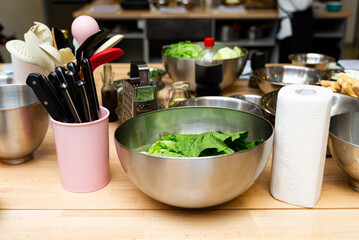 Kitchen Prep with Fresh Greens and Utensils. A well-equipped kitchen prep area with fresh greens in a large metal bowl, various cooking utensils, and bowls on a wooden counter.