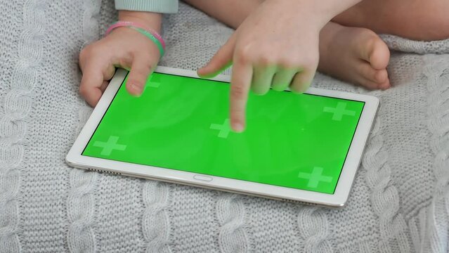 A tablet with a green chrome kay screen lies on a gray knitted plaid, a little girl touches the display with her finger. Bare feet. Advertising an app, video games.