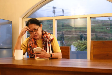 A middle-aged woman checks her smartphone while a book and a white of cup are in front of her