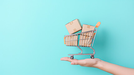 A person's hand holding a mini shopping cart filled with parcels against a turquoise background, representing online shopping and delivery.