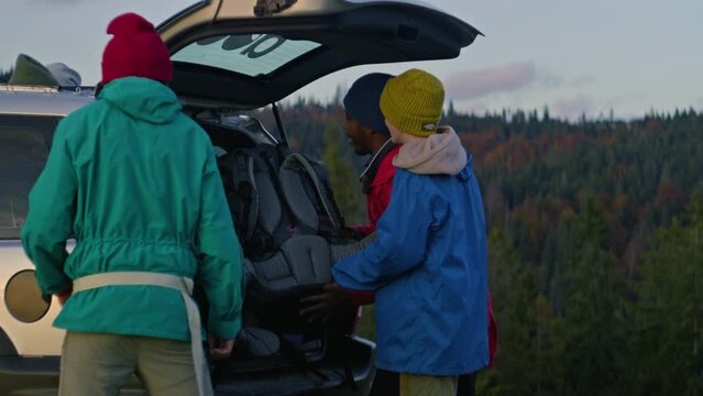 Group of diverse travelers pack their luggage and backpacks into the trunk of car preparing for hike. Tourist family or hiking buddies during road trip on vacation in the mountains. Slow motion.