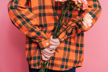 A boy in a checkered shirt holds a bouquet of flowers behind his back against a pink background.