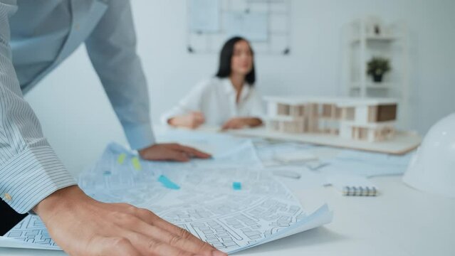 Skilled caucasian interior designer working with house model while discussing with asian architect engineer about house plan on meeting table with blueprint and house model scatter around. Immaculate.