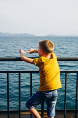 A boy in a yellow T-shirt stands on the deck of the ferry and looks at the sea. The child looks into the distance
