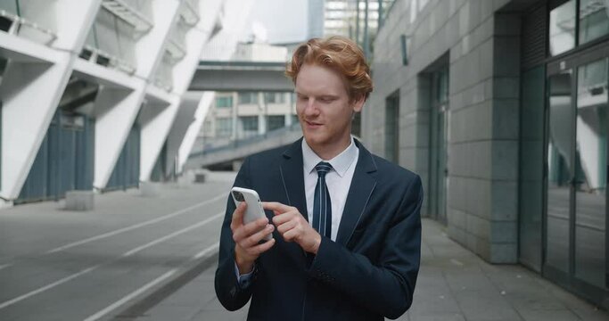 Office worker in formal suit on street surfs social networks. Man took break near office building to distract himself with phone. Worker in office suit is looking at news on phone with smile.