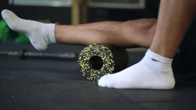 Close-up. Live camera. A young man does shin rolling using a massage roller while sitting on the floor in the gym. Myofascial release using a massage roller