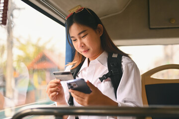 Happy young holding credit card and using mobile phone while sitting inside public bus transport