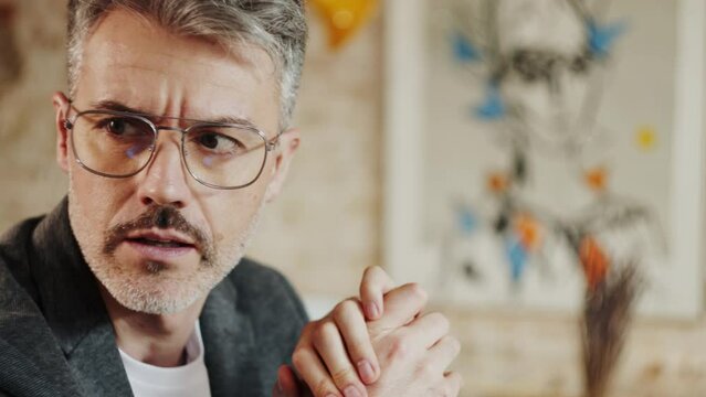 handsome businessman with bristle and moustache in eyewear talking to his business colleague with hands crossed. He is nodding and smiling. Blurred painted in the background.