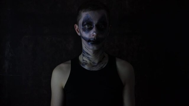 Young Caucasian man with a scary dark Halloween clown makeup costume standing in front of a black background