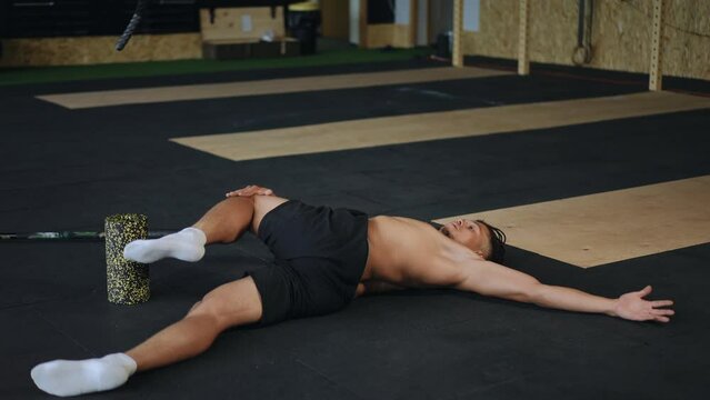 A man lies with his arms outstretched on the floor in the gym and performs crunches, trying to touch the floor with his knees without lifting his back
