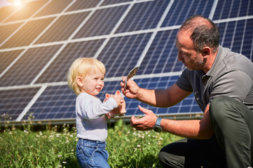 Father and his happy son saving money in piggy bank on background of solar panels. Man and his cute child putting some cash to piggy bank. Concept of saving money, investment in renewable energy.