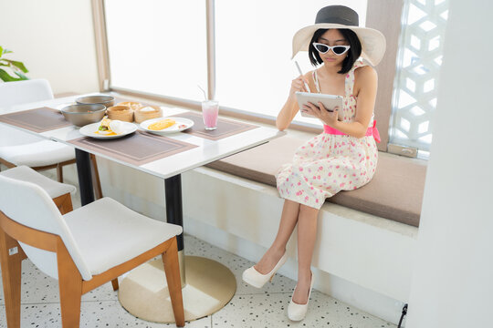 Full body image of Asian female tourist, wearing glasses, dress and hat, doing business with a laptop in a hotel restaurant. On the table were Asian, European breakfasts, desserts and drinks.