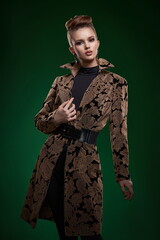 A young girl with exquisite makeup and a high stylish hairstyle with a gold decoration on her neck in a stylish coat, with well-groomed hands, poses in the studio on a green background