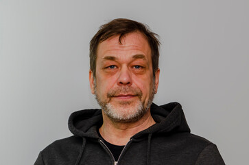 An elderly 50-55 year old man with a beard wearing a black hoodie looks into the camera against a light colored background.