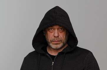 An elderly man 50-55 years old with a beard and wearing a black hooded hoodie looks into the camera against a gray background.