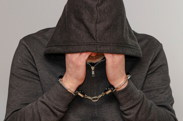 Hooded man handcuffed against a light-colored background, hands resting against his face.
