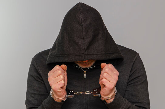 Hooded man handcuffed against a light colored background, hiding his face.