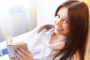 Cute brunette girl is reading small book of poetry in bedroom in morning. Young pregnant woman in the maternity hospital with a book of tips on parenting. A sick and recovering lady