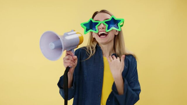Cheerful woman in large stars glasses talking in loudspeaker and gesturing with hands. Excited female making loud announcement in megaphone about huge discounts and laughing on yellow background.