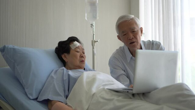 Asian senior couple talking and looking and business through computer in hospital room while injure