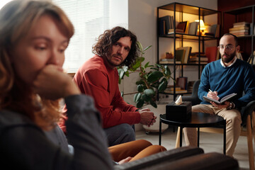 Selective focus shot of Caucasian man sitting on couch in psychologists office looking at his depressed wife
