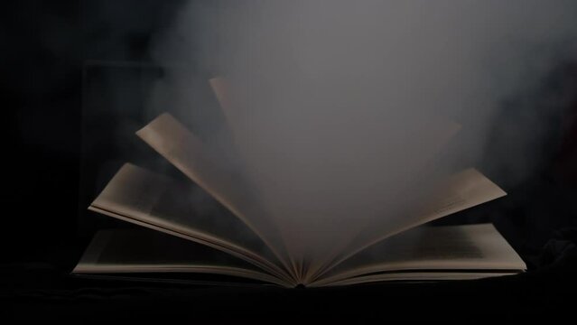 Old mystical book with open pages with magic smoke on black background. Enchanted book with magic light in the dark. Open book lies on the table, illuminated by beam of light among the falling smoke.