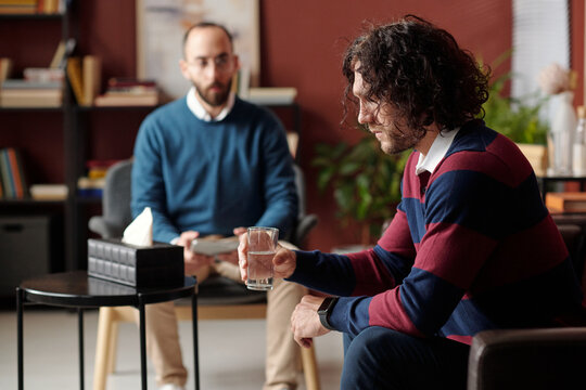 Selective focus shot of young man with messy curly hair suffering depression holding glass of water having psychotherapy session