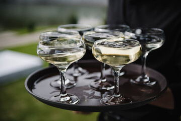 The waiter serving champagne on tray. Waiter holding glasses in the backyard. Tray of glasses of summer sparkling wine for guests at wedding reception. Man brings full glasses of champagne on a tray