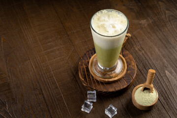Ice matcha latte in a tall glass with wooden coaster and matcha green tea powder in wood table