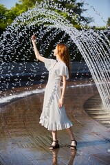 A beautiful young girl in a light summer polka dot dress stands in the park near the fountain. The...