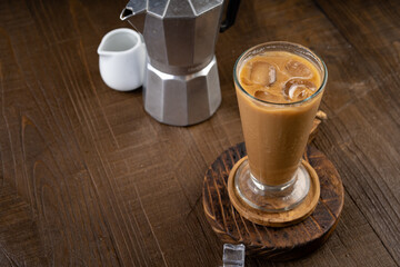 Ice coffee latte in a tall glass with wooden coaster and moka pot coffee maker