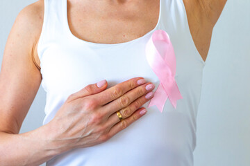 Shot of the woman against the white wall in the white top with pink ribbon, as a symbol of a breast cancer awareness, performing self examination of the breasts, looking for abnormalities. Concept