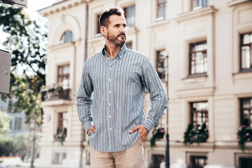 Fototapeta premium Portrait of handsome confident stylish hipster lambersexual model. Sexy modern man dressed in blue shirt and trousers. Fashion male posing on street background in Europe city at sunset