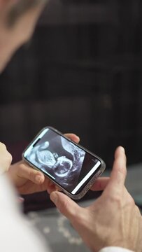 Faceless Pregnant Couple on Sofa Watching And Pointing a Phone with Ultrasound Image. Unrecognizable pregnant couple on sofa, woman showing ultrasound image on phone.