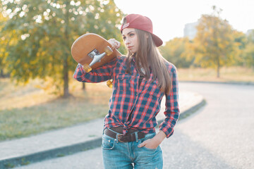 Cool hipster girl skater with a cap in a fashion plaid shirt and jeans holds a skateboard and stands in the park © alones