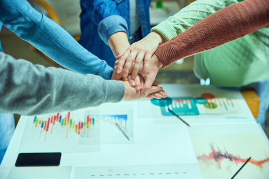 Hands of colleagues places on each other over a table with business graphs, symbolizing agreement and partnership. Start of new project and cooperation. Concept of business, teamwork, modern office