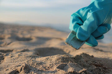 Close-up of hand with cleaning gloves picking up a small plastic from the sand on the beach with...
